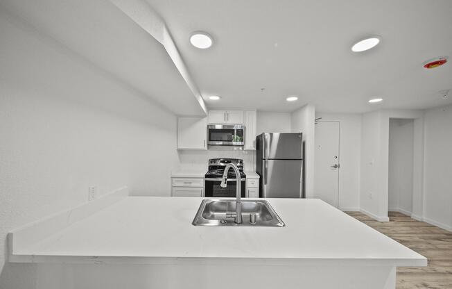 Modern kitchen with white cabinetry, stainless steel appliances including a fridge and stove, and a center island featuring a sink. The walls are painted white, and the flooring is light wood. Bright lighting from recessed fixtures enhances the spacious feel of the room.
