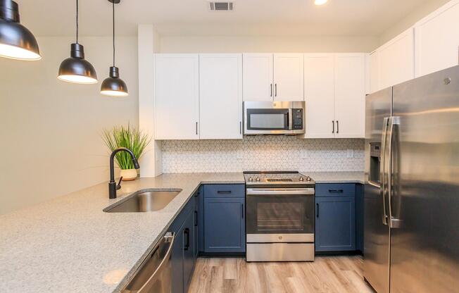 A modern kitchen featuring white and navy blue cabinetry, stainless steel appliances including a microwave and oven, and a large sink. The countertop is made of gray stone, and stylish pendant lights hang above. A decorative plant adds a touch of color to the space.