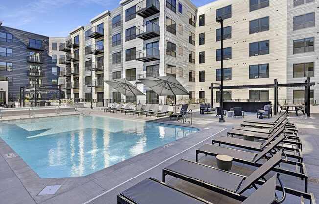 A large pool surrounded by sun loungers and umbrellas in front of apartment buildings.