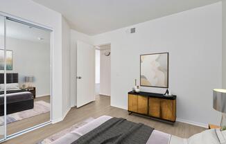 A modern bedroom featuring a neatly made bed with a patterned throw, a wooden dresser with decorative items, and a large mirror reflecting the room. The walls are painted white, and there are two doors leading to other spaces. Natural light fills the area, enhancing the contemporary decor.