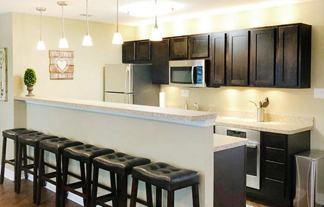 A modern kitchen with dark wooden cabinets, stainless steel appliances, and a granite countertop. There are three bar stools with black cushions at the kitchen island, and pendant lights hanging above it. A decorative wall piece is visible on the side.