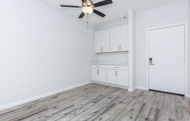 A spacious, empty room with light wood flooring, a ceiling fan, and white walls. It features white cabinetry along one wall and a door leading outside. Natural light enhances the airy feel of the space.
