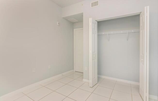 A bright, empty room with light gray walls and white tiled flooring. It features an open closet with a single shelf, and two doors leading into the room. The space is well-lit, creating a clean and minimalistic atmosphere. There are no furnishings or decorative elements present.