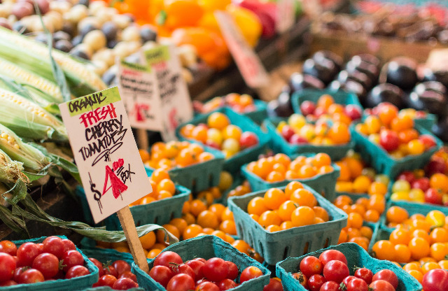 Stock photograph of produce at a farmer's market, featuring boxes of tomatoes, corn, potatoes, and peppers.