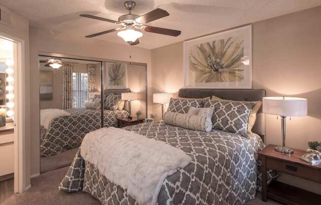 One and Two-Bedroom Apartments in Ahwatukee, AZ - Pacific Bay Club - Bedroom with Mirrored Closet Doors, Carpet Floors, Ceiling Fan, Cozy Bed, Stylish Decor, and Door to Bathroom with Wood-Style Plank