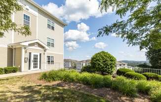 Exterior view of apartment building with beige siding and lush landscaping with view of additional buildings and panoramic view in background at Summit Place apartments in Methuen, MA.