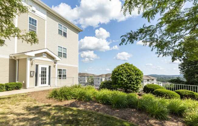 Exterior view of apartment building with beige siding and lush landscaping with view of additional buildings and panoramic view in background at Summit Place apartments in Methuen, MA.