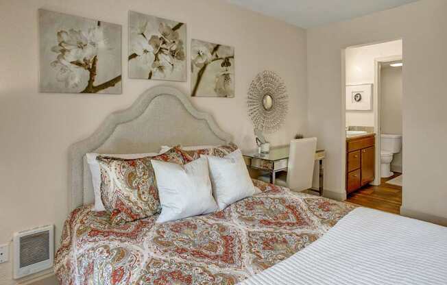 A welcoming bedroom here at Heritage Hills featuring a plush upholstered headboard, patterned bedding layered with neutral pillows, and coordinated floral wall art above the bed. The room includes soft carpeting, a mirrored vanity with chair, decorative wall mirror, and an open doorway leading to a bathroom with wood-style flooring and warm cabinetry.