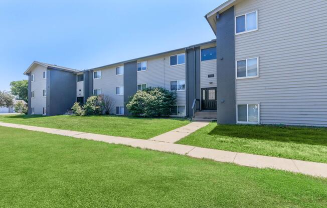 A modern, multi-unit residential building with gray siding, featuring multiple windows and a front walkway leading to the entrance. The lawn is well-maintained with lush green grass, and there are trees and shrubs planted around the exterior, under a clear blue sky.