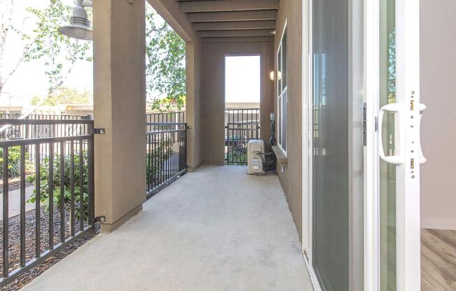 A covered patio area with concrete flooring, partially enclosed by a black railing. There are potted plants nearby, and a window with a door leading inside. The scene is well-lit, suggesting daytime, and features a clear view of the outdoors.
