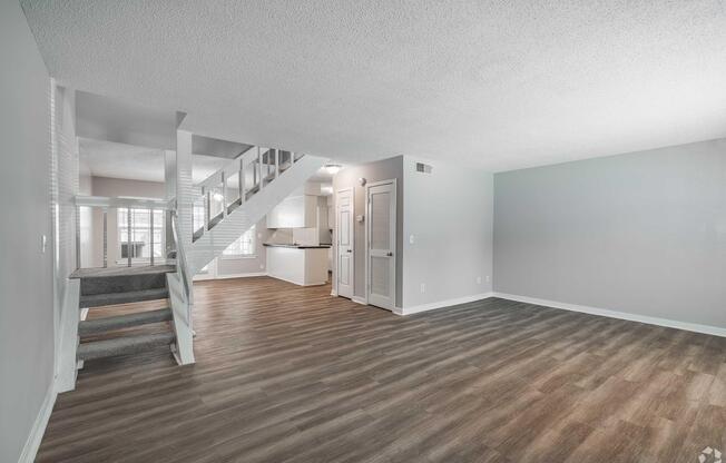 Spacious living area featuring a light gray color scheme, hardwood-style flooring, and a staircase leading to an upper level. The open floor plan connects to a modern kitchen visible in the background. Natural light filters through large windows, creating a bright and inviting atmosphere.