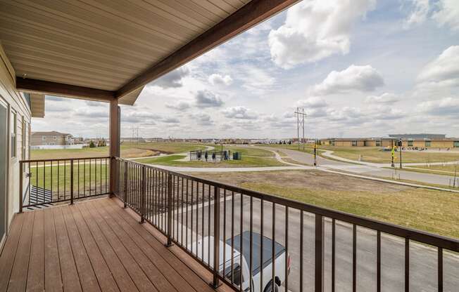 A balcony with a wooden floor and a metal railing overlooks a parking lot and a road.