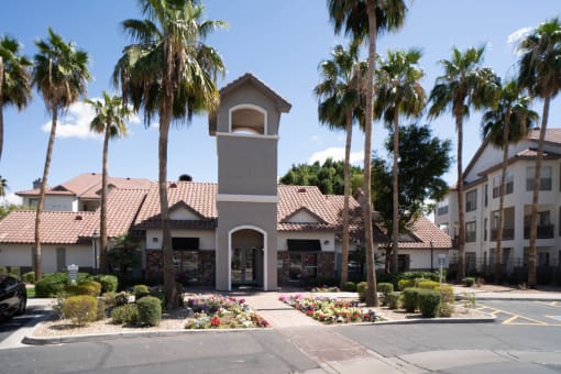 Apartments In Phoenix, AZ - Exterior View Of Leasing Office Surrounded By Palm Trees, Flowers, Lush Green Bushes, And Adjacent Apartment Buildings