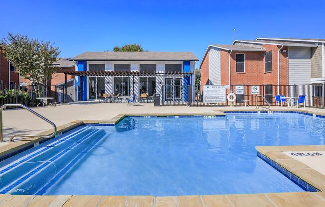 A clear blue swimming pool with a surrounding deck and lounge chairs, adjacent to a two-story building with large windows. The scene is set in a sunny outdoor area with trees in the background and a fence enclosing the pool space.