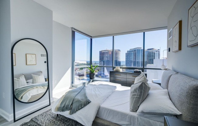 A modern bedroom featuring a plush bed with grey bedding, a large floor mirror, and a panoramic view of the city skyline through floor-to-ceiling windows