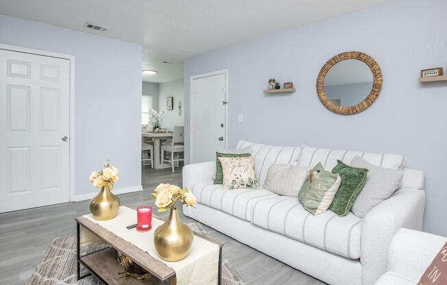 A cozy living room featuring a light gray sofa adorned with decorative pillows, two gold-colored vases with yellow flowers on a coffee table, and a circular mirror on the wall. A white door leads to another room, and a dining area with a light wooden table and chairs can be seen in the background.
