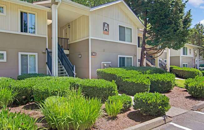 A well-maintained apartment building exterior here at Asbury Park featuring light-toned siding with contrasting trim, covered staircases with railings, and large windows, surrounded by neatly trimmed hedges, landscaped greenery, and mature trees, creating a clean, residential setting with clearly marked building signage and walkable paths.