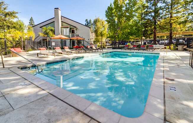 A large outdoor swimming pool surrounded by a patio area with chairs and a building in the background.