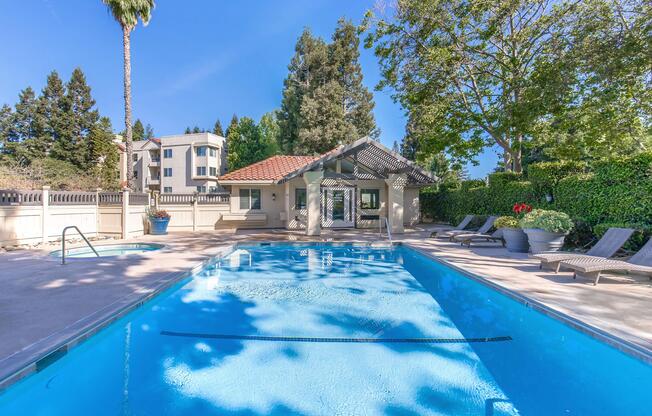 A clear blue swimming pool surrounded by lounge chairs and potted plants. In the background, there are trees and a building with a Mediterranean-style roof. The scene is bright and sunny, creating a relaxing outdoor atmosphere.