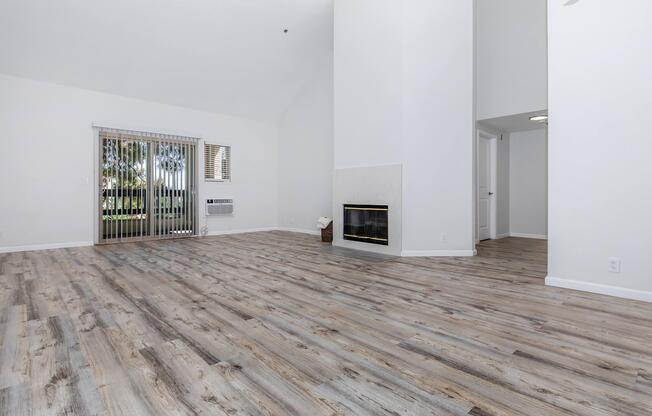 A spacious living room with light-colored walls and wood-style flooring. It features a white fireplace on one side, tall ceilings, and a large window letting in natural light. Vertical blinds cover the window, and there's an air conditioning unit mounted on the wall. The room is empty, creating an open and inviting atmosphere.