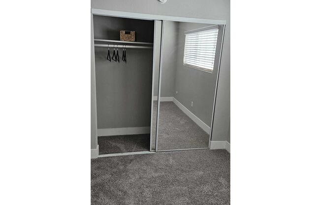 A cozy room featuring a mirrored closet with sliding doors. Inside the closet, there are several hangers and a small basket on a shelf. The floor is covered with plush, light-colored carpet, and there's a window with blinds allowing natural light to filter in. The walls are painted a soft gray.
