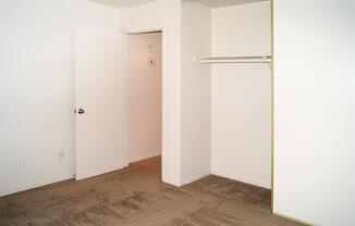 Empty room with beige carpet, white walls, and a closet without doors. A light-colored door leads to another space, and the room has minimal furnishings. There are no decorations or items visible, creating a spacious and clean appearance.