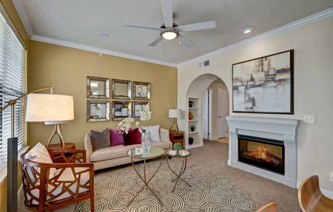 A stylish living room here at Tuscany at Faudree with plush carpet, a comfortable sofa accented by pillows, and a round patterned area rug. A built-in fireplace creates a cozy focal point beneath wall art, while a ceiling fan, table lamps, and arched doorway add warmth, light, and an inviting, open feel.