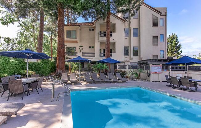 A serene outdoor pool area with a clear blue pool surrounded by lounge chairs and umbrellas. In the background, a multi-story building with balconies and tall trees adds to the tranquil atmosphere. Perfect for relaxation and leisure on a sunny day.