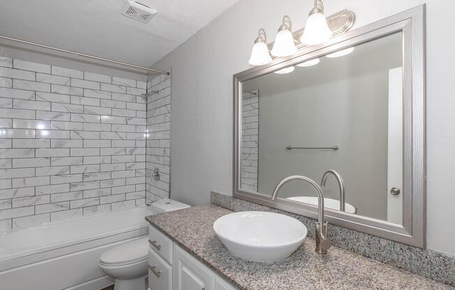 A modern bathroom featuring a bathtub with a tiled shower, a granite countertop with a stylish round sink, and a large mirror above the sink. Three wall-mounted light fixtures illuminate the space, which has neutral-colored walls and an organized aesthetic.
