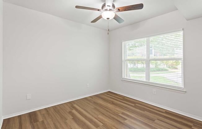 A spacious, empty room featuring light-colored walls and a wooden floor. A ceiling fan with wooden blades is installed above, and a large window with blinds allows natural light to illuminate the space. The room has a clean, minimalist design, suitable for various uses.