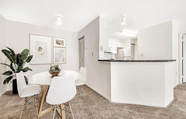 Virtually staged dining area with carpet, white table with chairs, indoor plants overlooking kitchen with breakfast bar