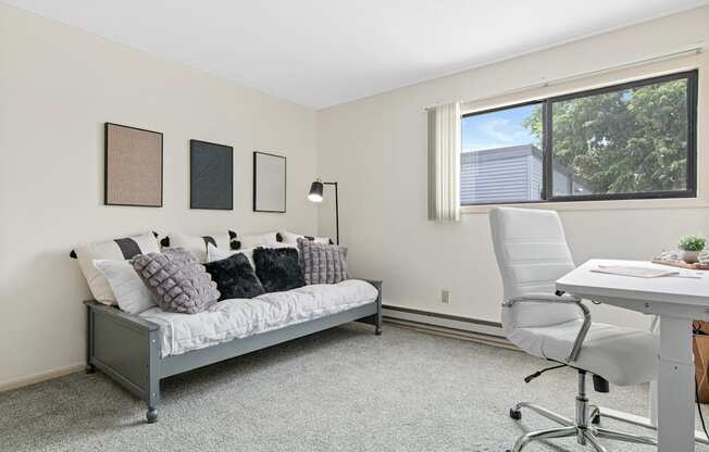 A room with a grey sofa, a white chair, and a desk with a plant on it.
