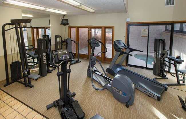 A bright fitness center here at Eagle Ridge showcasing a well-equipped workout space with treadmills, an elliptical, strength training machines, and free weights arranged on carpeted flooring, with large windows and glass doors providing natural light and views into the adjacent indoor pool area.