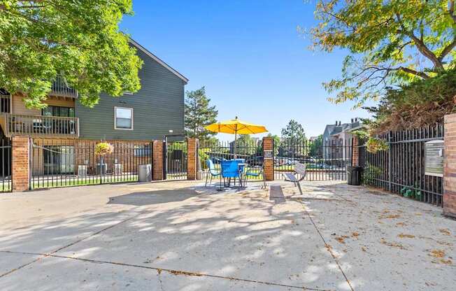 A playground with a yellow umbrella and a blue slide is surrounded by a fence and a building.