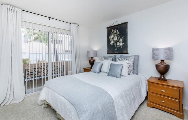 A cozy bedroom featuring a queen-sized bed with light gray bedding and decorative pillows. On either side of the bed are wooden nightstands with lamps. Large windows with vertical blinds allow natural light in, and a floral artwork hangs above the bed, adding a touch of elegance. Soft carpet covers the floor.