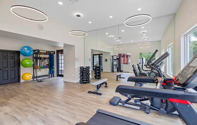 Fitness center with mirrored wall, strength training equipment, and treadmills. Flat screen televisions above large windows with view of community areas