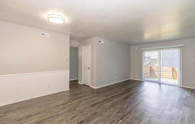 A spacious, empty living room with light-colored walls and a wood-like floor. There is a ceiling light fixture, a sliding glass door leading to a small patio or balcony, and a white paneling accent on the lower wall. The room has a clean and modern appearance, ideal for furniture arrangement.