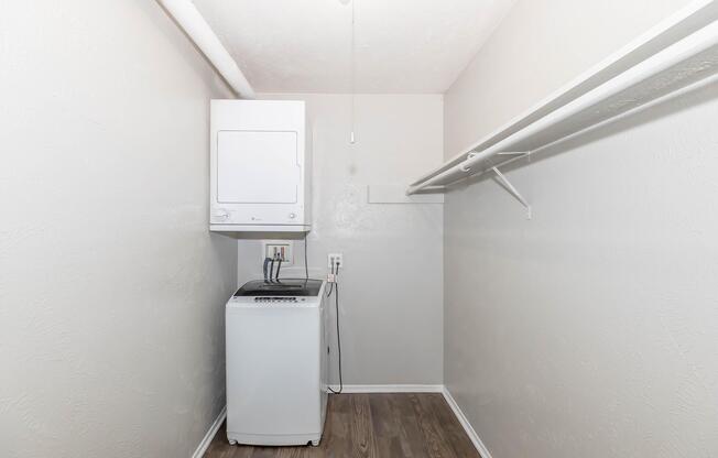 A laundry room featuring a stacked washer and dryer unit alongside a clothes rack. The walls are painted light gray, and the floor has a wood-like appearance. The space is well-lit with a ceiling light, providing a clean and modern look.