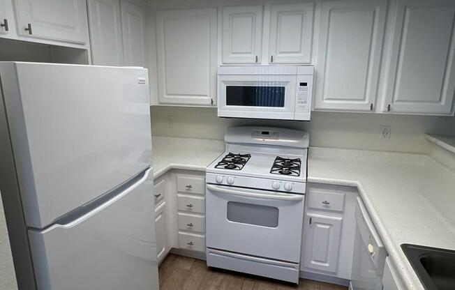 A modern kitchen featuring white cabinetry, a white refrigerator, a built-in microwave above a gas stove, and a light-colored countertop. The layout is compact with ample storage space and a clean, bright aesthetic. The flooring is a light wood finish, complementing the overall design.