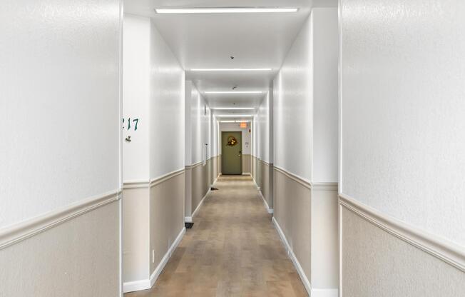 A well-lit corridor with white walls and light-colored flooring, featuring a decorative wall element at the end. Doors line either side, with a visible door marked "217." The lighting is uniform, creating a spacious and clean atmosphere.