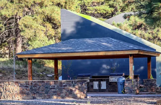 A wooden pavilion with a stone wall and a green roof is surrounded by trees.