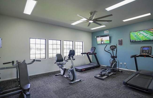 A gym room with a row of treadmills, a fan, and a television showing a baseball game.