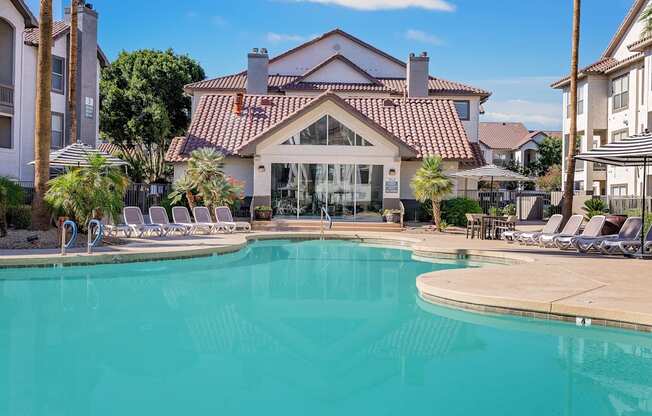 A large swimming pool in front of a house with a patio and lounge chairs.