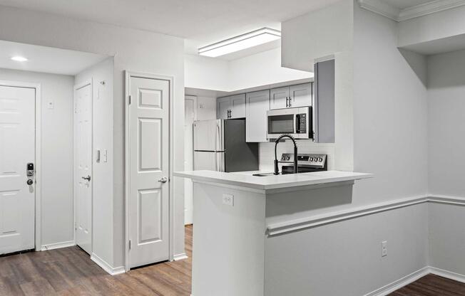 Modern kitchen and living area, featuring an open layout with white walls, a small breakfast bar, stainless steel appliances including a refrigerator and microwave, and wood-style flooring. The entrance door is visible on the left side, and there is ample natural light from the ceiling fixtures.