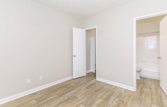 A minimalist room with light gray walls and a wooden floor. The space features a white door leading to a small closet, another white door that opens to a bathroom, and an overall clean and bright appearance, ideal for personal or rental use.