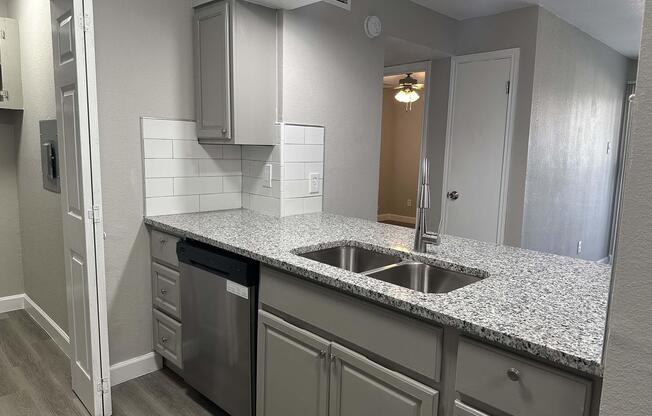 A modern kitchen featuring light gray cabinetry, a stainless steel dishwasher, and a double sink with a sleek faucet. The countertop is made of speckled granite, and there are white subway tiles as a backsplash. Soft lighting enhances the inviting atmosphere, with an open doorway leading to another room in the background.