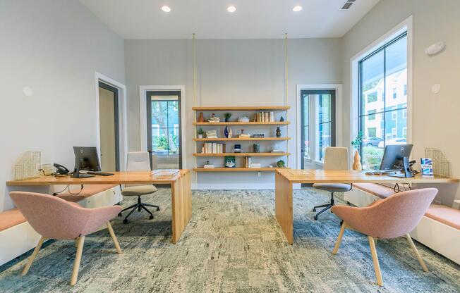 A bright and modern office space featuring two wooden desks with computers and telephones. There are pink ergonomic chairs, a bookshelf filled with decorative items, and large windows allowing natural light to fill the room. The floor is covered with light-colored carpet, creating a cozy atmosphere.