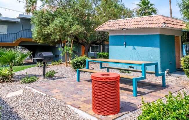 Fifteen 50 apartments Las Vegas courtyard with modern blue picnic table, market lights, red trash can, and barbecue next to small blue structure.