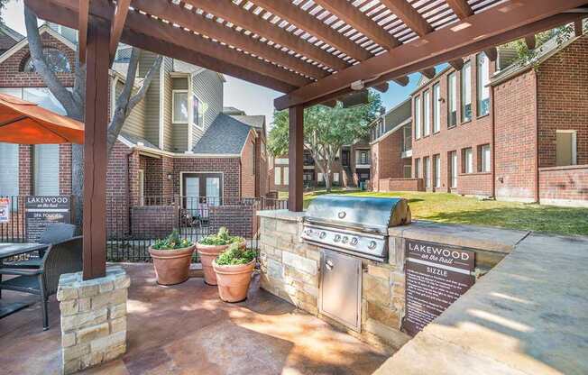 A covered outdoor grilling station here at Lakewood on the Trail with a stainless steel grill, stone counter, pergola shade, and nearby patio seating, creating a convenient and inviting cookout area.