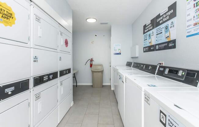 A clean and well-lit laundry room featuring several stacked washing machines and dryers against the wall. There is a trash bin and a fire extinguisher visible. Informational signs are posted on the wall, providing instructions and services related to laundry. The floor is tiled and the space looks organized.